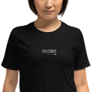 「STAY CURIOUS」刺繍Tシャツ ブラック｜胸元の白い刺繍のアップ。文字からほどけた糸を猫が引っぱっている